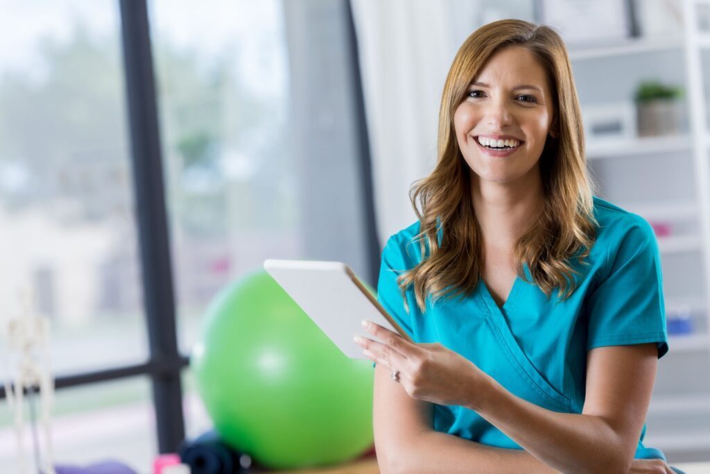 Physical therapist wearing scrubs and holding a tablet while smiling at the camera