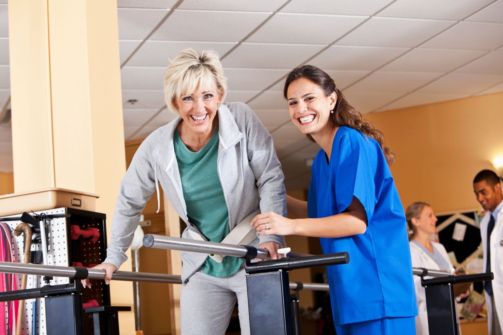 A physical therapist and her patient smiling at the camera as the therapist helps her patient walk