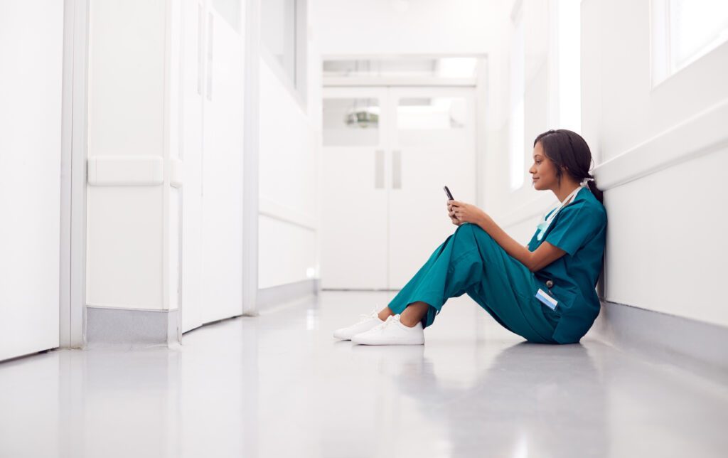 Female Nurse Wearing Scrubs Sitting On Floor In Hospital Corridor Using Mobile Phone